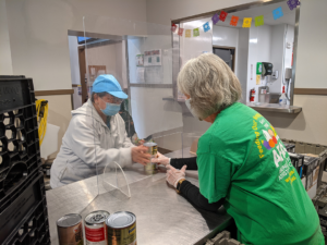 AFAC volunteer in green shirt handing canned goods to a woman behind a plexiglass barrier